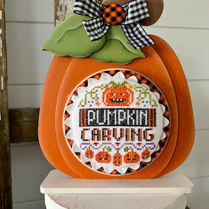 May include: A wooden pumpkin decoration with a black and white checkered bow and a cross-stitched design that says "Pumpkin Carving".