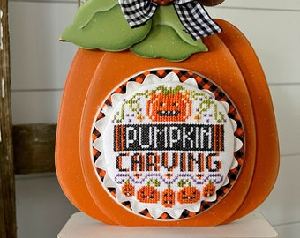 May include: A wooden pumpkin decoration with a black and white checkered bow and a cross-stitched design that says "Pumpkin Carving".
