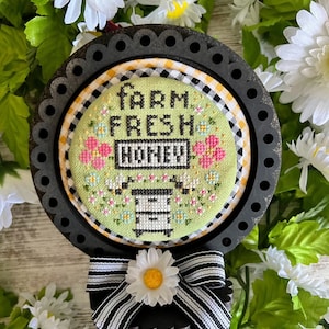 Pode incluir: Um aro de bordado enquadrado em preto com um desenho de ponto cruz verde e branco que apresenta uma colmeia e o texto "Farm Fresh Honey". O aro é decorado com uma fita listrada preta e branca e uma margarida branca.