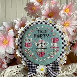 May include: A teal blue cross-stitch embroidery hoop with a black and white checkered fabric backing. The embroidery depicts a teapot and teacups with the text "Tea Party". The hoop is decorated with a white daisy and a black and white checkered bow.