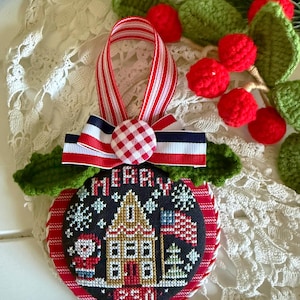 May include: A round Christmas ornament featuring a cross-stitch design of a house, a flag, and a snowman on a black background with the text Merry 250. It is accented with a red and white striped ribbon, a gingham button, and green crochet leaves.