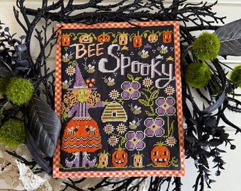 May include: A framed cross-stitch featuring a Halloween theme. The design includes a witch, pumpkins, flowers, and the words "Bee Spooky." The artwork is set against a black background with an orange and white gingham border.