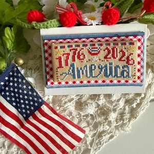 May include: A patriotic cross-stitch design with the text "1776 2026 America" in red and blue. The design is on a cream-colored background with a gingham border. An American flag and decorative elements complete the scene.