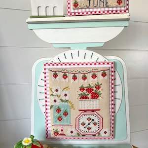 May include: A decorative shelf with a strawberry theme. It features a wooden strawberry basket, a cross-stitch piece with the word "JUNE", and a larger cross-stitch design with strawberries and flowers. A ceramic strawberry and wooden beads complete the display.