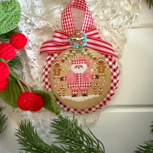 May include: A round Christmas ornament featuring a cross-stitch design of a Santa Claus in a pink suit flanked by two gingerbread figures. The ornament is framed by a red and white gingham fabric border and topped with a matching ribbon bow.