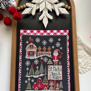 Può includere: Un ricamo a punto croce con un camioncino rosso, un fienile e Babbo Natale con le renne. Il disegno è su tessuto nero con un bordo a quadri rossi e bianchi. Il testo "St. Nick's Tree Farm" è incluso nel disegno.
