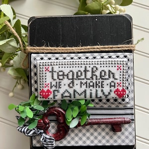May include: Black wooden sign with a white and black checkered background. A cross-stitched piece with red hearts reads "Together we make a family". A red metal key with a black and white ribbon is attached to the sign.