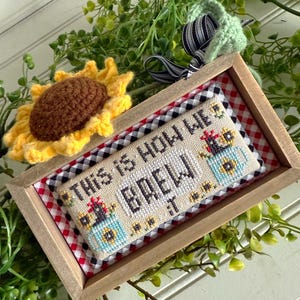 May include: A framed cross-stitch with a red and white checkered background. The cross-stitch reads "This is how we brew it" and features a cartoon image of a rooster holding a blue pitcher with sunflowers.