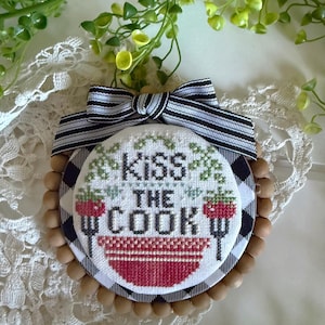 以下が含まれることがあります： 黒い文字で「Kiss THE COOK」と刺繍された円形のクロスステッチ壁掛け。フォークに乗った2つの赤いイチゴと赤いボウルが描かれています。黒と白のチェック柄の縁取りと、天然木のビーズで装飾されています。