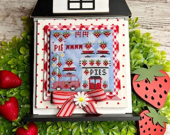 May include: A cross-stitch embroidery piece framed in a black house-shaped frame. The embroidery depicts a red car with a sign that reads "Pie Wagon Pies". The embroidery is surrounded by red and white gingham fabric and red polka dot fabric. The frame is decorated with a red and white striped ribbon and a white daisy.
