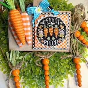 May include: A wooden sign with a black and white checkered background and a black circle with the text "Carrot Patch" and a picture of carrots and bunnies. The sign is surrounded by a green wreath with orange carrot-shaped beads hanging from it.