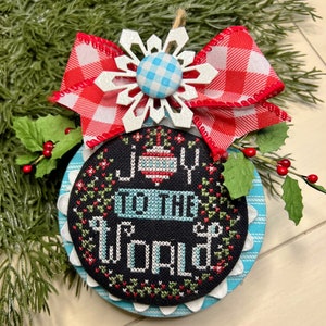 May include: A blue and white Christmas ornament with a red and white checkered bow. The ornament is embroidered with the words "Joy to the World".
