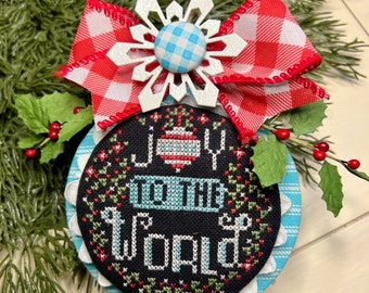 May include: A blue and white Christmas ornament with a red and white checkered bow. The ornament is embroidered with the words "Joy to the World".
