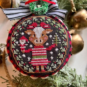 Puede incluir: Un adorno navideño redondo con fondo rojo y negro a cuadros. El adorno presenta un diseño de punto de cruz de una vaca con un gorro de Papá Noel y sosteniendo un bastón de caramelo. La vaca está rodeada de árboles verdes y copos de nieve.