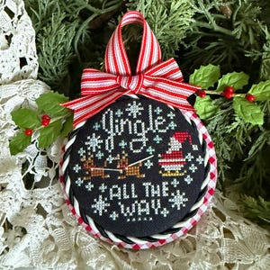 May include: A round, black Christmas ornament with the words "Jingle All The Way" in white cross-stitch. The ornament features a Santa Claus and reindeer design, a red and white striped bow, and a red and white gingham border.
