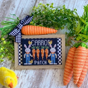 May include: A framed cross-stitch design featuring two bunnies holding carrots with the text "Carrot Patch". The frame is wooden with a black and white checkered background. The image is surrounded by green foliage and orange carrots.