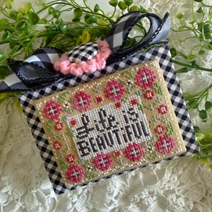 May include: A cross-stitched piece with the words "Life is Beautiful" surrounded by pink flowers and green leaves. The piece is set against a tan background and framed with a black and white gingham pattern. A pink flower and gingham ribbon complete the design.