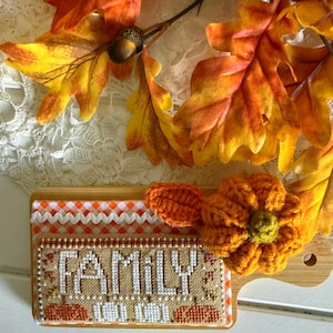 May include: A rectangular wooden board featuring a cross-stitched fabric insert with the word FAMILY in white letters, flanked by small orange pumpkins. A crocheted orange pumpkin with a green center is attached to the side, accented by autumn leaves.