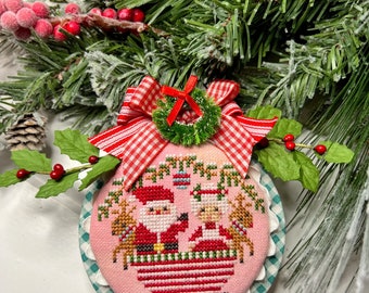 May include: A pink and white Christmas ornament with a cross-stitch design of Santa Claus and Mrs. Claus riding in a sleigh pulled by reindeer. The ornament is decorated with a red and white gingham bow and a green wreath.