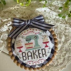 May include: A circular cross-stitch ornament featuring a light green stand mixer, two cakes on red stands, and the word BAKER in black letters. The piece is framed by a black and white buffalo check border, wooden beads, and a striped fabric bow.