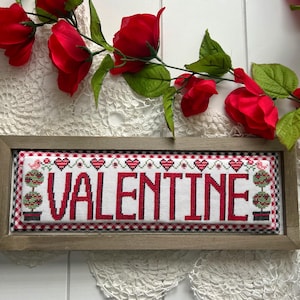 May include: A framed cross-stitch with a red and white checkered border. The cross-stitch features the word "VALENTINE" in red lettering with a red heart design above and below the word. The cross-stitch is surrounded by a wooden frame.