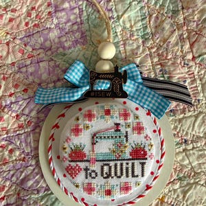 Könnte beinhalten: Runder Zieranhänger mit Kreuzstichmuster. Er zeigt eine Nähmaschine, Erdbeeren und den Schriftzug "to QUILT". Er hat einen rot-weißen Seilrand, eine blau-weiß karierte Schleife und einen Holzperlenaufhänger. Der Text "#ISEW" ist ebenfalls sichtbar.