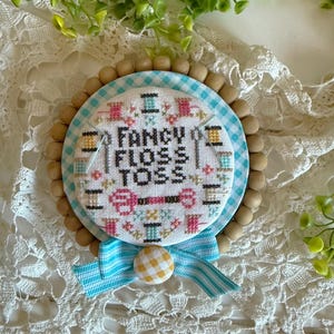 Puede incluir: Un bordado de punto de cruz circular con el texto "FANCY FLOSS TOSS", rodeado de cuentas de madera. El diseño presenta carretes, tijeras y flores en rosa, amarillo y azul. Una cinta y un botón de cuadros azules y blancos acentúan la pieza.