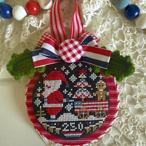 May include: A round fabric ornament featuring a cross-stitch design of Santa Claus, a Christmas tree, and a gingerbread man on a black background. The text 250 is stitched at the bottom. It is accented with a red and white striped ribbon, a gingham button, and green crocheted leaves.
