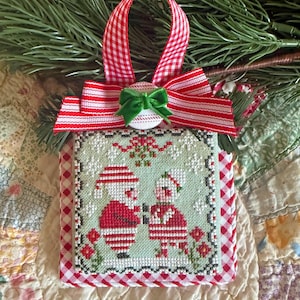 May include: A square cross-stitch ornament featuring a light green background with a Santa Claus and an elf figure. The ornament is framed by a red and white gingham border and topped with a red and white striped ribbon bow and a small green bow.