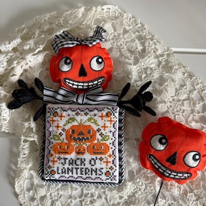 May include: A stuffed orange pumpkin figure with a black and white gingham bow, black felt arms, and a square embroidered panel featuring three pumpkins and the text JACK O' LANTERNS, accompanied by a separate orange pumpkin head on a black stick.