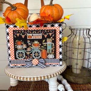 May include: A black and white checked tote bag with an embroidered design of a pumpkin patch, a scarecrow, and a bicycle with pumpkins. The bag is decorated with a black and white gingham ribbon and a white button. The bag is sitting on a white pedestal with a brown wicker basket and white beads in the background.