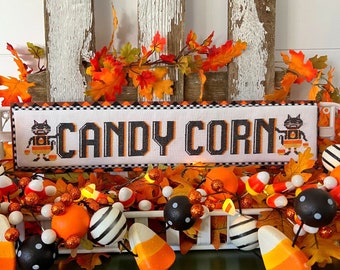 Pode incluir: Uma exibição de Halloween com um letreiro bordado em ponto cruz que diz "CANDY CORN" em preto e laranja. O letreiro apresenta gatos de desenho animado e fica acima de uma guirlanda de doces de milho, abóboras e folhas de outono. O fundo inclui uma estrutura de madeira branca desgastada.