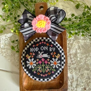 May include: A wooden serving board with a black and white gingham bow and a pink and yellow flower. A cross-stitched coaster with the words "HOP ON BY" and a bunny and floral design is attached to the board.