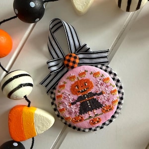May include: A pink, round, cross-stitched Halloween ornament featuring a scarecrow design. The ornament has a black and white gingham backing and a black and white striped ribbon. A garland of painted wooden beads and candy corn is in the background.