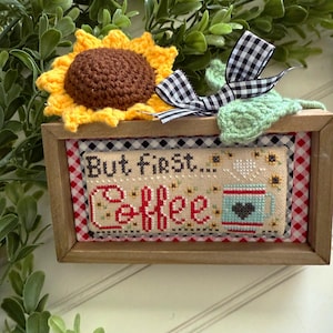 May include: A framed cross-stitch with a red and white gingham border. The cross-stitch reads "But first, coffee" and features a coffee cup with a heart on it.