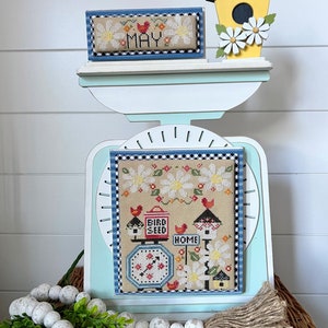 May include: A cross-stitch design featuring a birdhouse, a scale with the words "Bird Seed" and "Home", and daisies. The design is framed in blue and white checkered fabric and is displayed on a white scale with a wooden base. The scale is decorated with a yellow birdhouse with white daisies.
