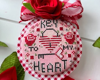 May include: A pink and white cross-stitch ornament with a red heart and the text "Key to my heart". The ornament is decorated with a red and white gingham ribbon and a red felt rose.