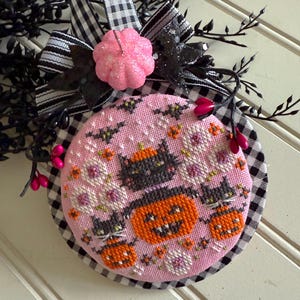 Peut inclure: Ornement de décoration d'Halloween rose et rond, avec un motif brodé de chats noirs, de citrouilles et de chauves-souris. L'ornement est bordé d'un tissu à carreaux noir et blanc et d'un ruban noir et blanc. Une citrouille rose à paillettes et des brins noirs complètent le look.