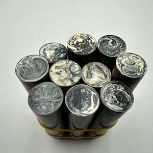 May include: A collection of nine cylindrical rods bundled together with a yellow rubber band. Each rod features a circular top surface with a marbled pattern of black, grey, and white swirls, while the sides of the rods are a solid dark colour.