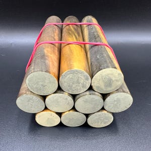 May include: A bundle of wooden dowels with varying shades of brown, gold, and gray, secured with a red rubber band. The ends of the dowels are a light gray color. The dowels are cylindrical in shape and appear to be made of wood.
