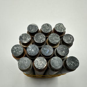 May include: A collection of fourteen cylindrical objects bundled together with a yellow rubber band. Each cylinder features a dark brown body and a circular top end with a marbled pattern in shades of black, grey, and white, creating a swirling effect.