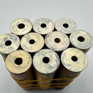 Puede incluir: Un grupo de nueve objetos cilíndricos dispuestos en conjunto y asegurados con una banda elástica amarilla. Cada cilindro presenta una superficie superior plana con un orificio circular central y un patrón jaspeado en tonos crema, blanco y gris.