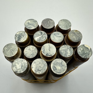 May include: A collection of fourteen cylindrical wooden objects arranged in a cluster and secured with a yellow rubber band. Each object features a flat, circular top surface with a marbled pattern in shades of white, grey, and black.