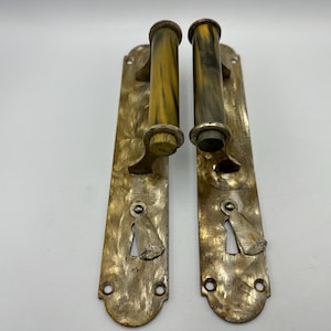 May include: Two matching door handles with long, rectangular, weathered brass backplates featuring a mottled finish. Each handle has a cylindrical grip with a marbled yellow and black pattern, a keyhole cover, and mounting holes at the ends.