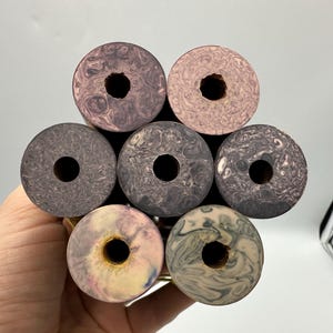 Puede incluir: Una colección de siete tapones decorativos para botellas con un diseño circular y un agujero en el centro. Los tapones presentan una variedad de colores, incluyendo rosa, gris y blanco, con un patrón arremolinado. Los tapones se mantienen unidos.