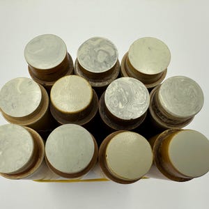 May include: An overhead shot of a collection of cylindrical bottle stoppers. Each stopper has a light-coloured, circular top with a marbled or solid appearance. The stoppers are brown and beige, arranged closely together.
