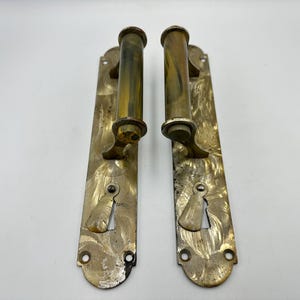 Puede incluir: Dos manijas de puerta de latón a juego con placas traseras rectangulares largas. Cada placa presenta una textura de metal cepillado con remolinos y un corte para cerradura en la parte inferior. Los mangos cilíndricos están montados horizontalmente.