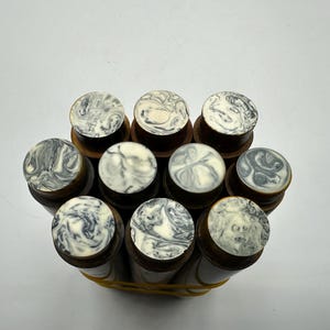 May include: A collection of nine cylindrical wooden objects bundled together with a yellow rubber band. Each object features a circular top inlay with a marbled pattern of black, grey, and white swirls, contrasting with the dark brown wooden base.