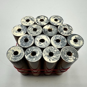 May include: A collection of fourteen cylindrical wooden pen blanks bundled together with a red rubber band. Each blank features a dark brown body and a circular top end with a marbled black and white resin pattern and a centred hole.