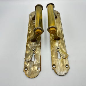 May include: Two matching door handles with long, rectangular brass backplates featuring a hammered texture. Each handle has a cylindrical, translucent yellow and brown marbled grip. A keyhole with a small, pivoting metal cover is located below.